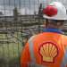 Shell worker with tabard emblazoned with Shell's logo looks out through a fence at oil and gas infrastructure Nigeria