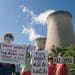 Climate protesters stand under enormous gas flue stacks at Drax power station near Selby, Yorkshire. They hold placards calling out the company's complicity. They read: "Wood pellets cause lung disease and cancer in local communities", "Trees support wildlife habitats" and "Axe Drax".