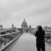 Grayscale image of St Paul's Cathedral in the background. Seven women/non-binary people stand nude with their backs to a photographer, in a line across Millennium Bridge photoshoot