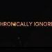 'Chronically Ignored' in orange text over a black background with a few small lights running down the ceiling of a pitch-black tunnel ME/CFS
