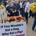 Activist with an apron reading 'End Speciesism" stands over a barbecue gridle with corn on the cob and a visually realistic dog and a banner reading "If you wouldn't eat a dog, why eat a pig? Please, go vegan!" People queuing to go into Meatopia festival.