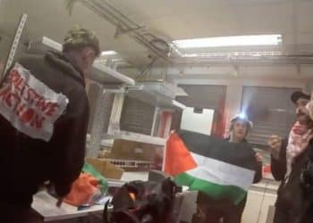 Palestine Action activists inside Elbit's Ulm factory holding a Palestine flag and a Palestine Action banner on one activist's back damaging equipment.