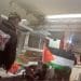 Palestine Action activists inside Elbit's Ulm factory holding a Palestine flag and a Palestine Action banner on one activist's back damaging equipment.