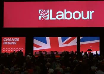 Labour Party conference stage from 2024, with crowds filtering in. Labour's logo is projected on a red backdrop. The slogan "Change begins" features on a red screen below next to a Union Jack on a screen beside it Israel