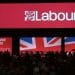 Labour Party conference stage from 2024, with crowds filtering in. Labour's logo is projected on a red backdrop. The slogan "Change begins" features on a red screen below next to a Union Jack on a screen beside it Israel