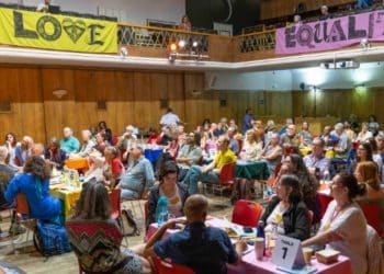 A large group of people sit together at tables in a room with "Love" and "Equality" banners above, for a people's assembly Your Party