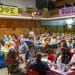 A large group of people sit together at tables in a room with "Love" and "Equality" banners above, for a people's assembly Your Party