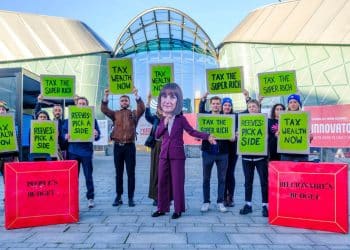 A figure wearing a large Rachel Reeves head stood between two oversized red Budget briefcase boxes, one labelled “billionaire budget”, the other “people’s budget”, surrounded by campaigners holding banners and placards reading “Tax the super-rich”, “Tax wealth now” and “Reeves: Pick a side” Labour conference