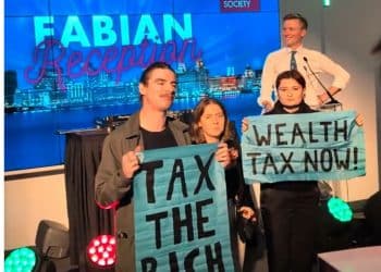 Torsten Bell on stage at the Fabian Reception at Labour's conference, as three activists stand in front of him holding placards reading "Tax the rich" and "Wealth tax now!"