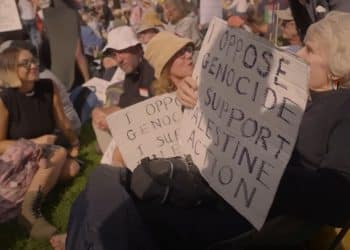 Protesters sit together holding "I oppose genocide. I support Palestine Action" placards, including an elderly woman with Palestine flag earrings Defend Our Juries
