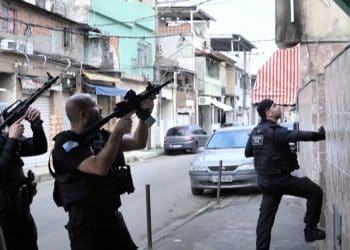 Three armed Rio de Janeiro police. Two point their guns up at a residence in the city's favelas, while one attempts to break down the door.